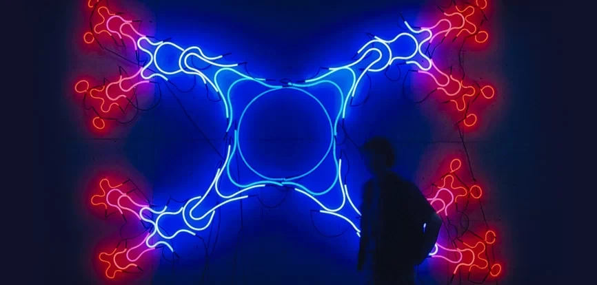 A large-scale neon wall installation with a symmetrical cross-shaped design in blue and red neon tubes, with a silhouetted figure standing in front of the glowing artwork in a darkened gallery.